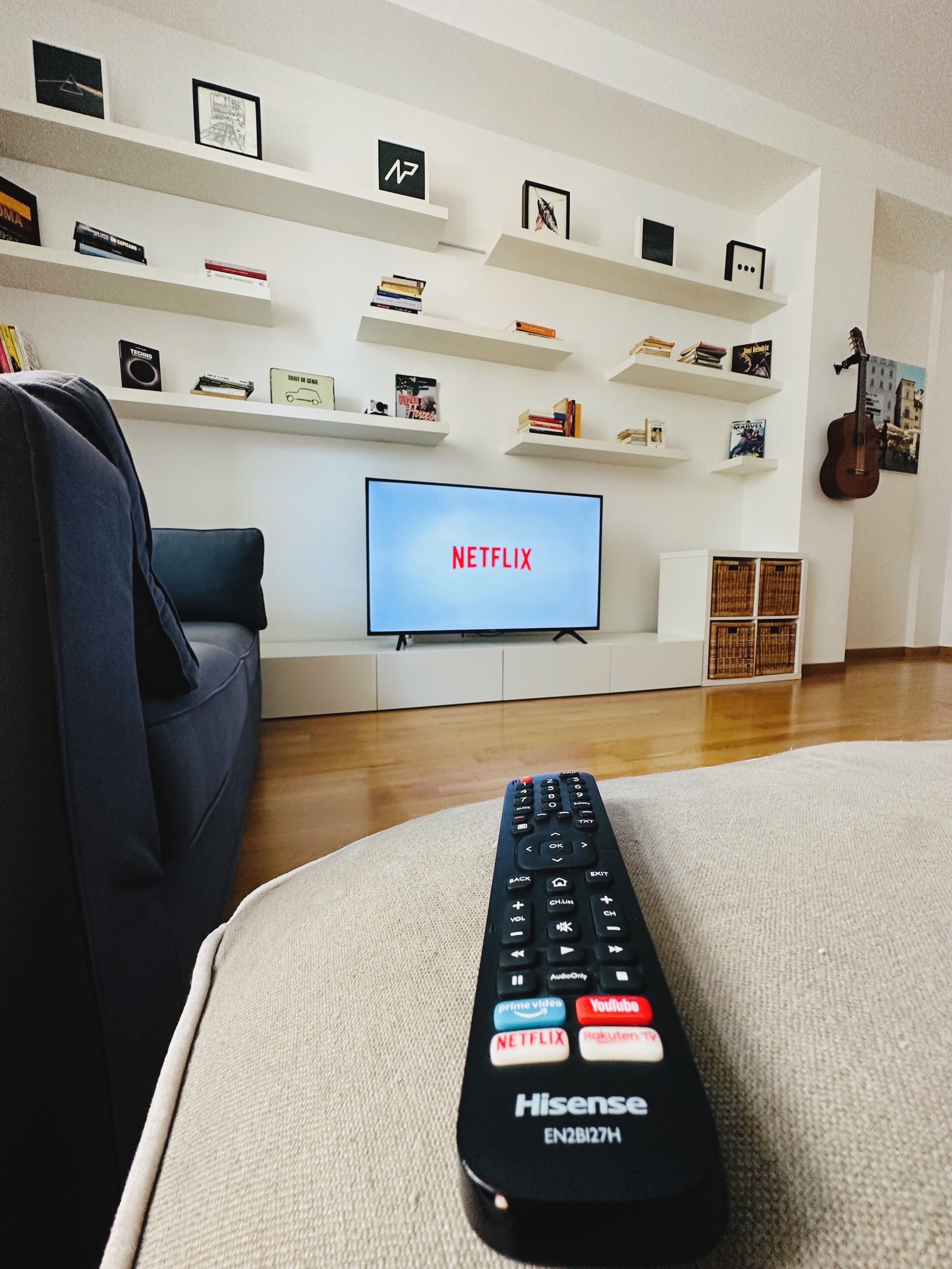 A living room with a television displaying 'NETFLIX' on the screen, a remote control on a couch, and shelves with books and decorative items.