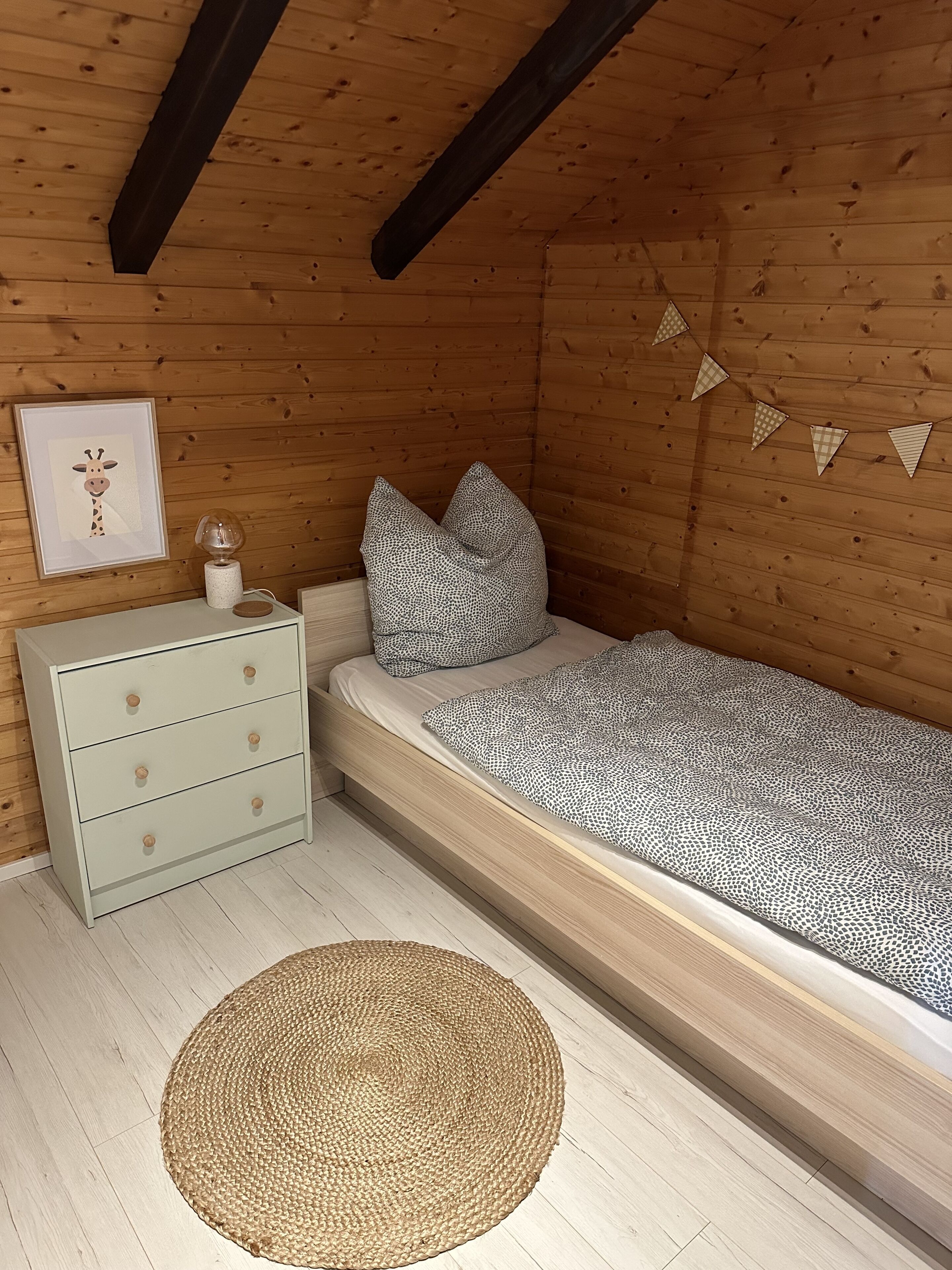 Ein Schlafzimmer mit Holzboden, einem Bett mit gemusterten Betttextilien, einem Nachttisch mit Schubladen, einem gerahmten Bild und einer dekorativen Kette aus Dreiecken.