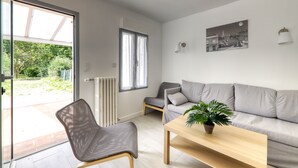 Een moderne woonkamer met een grijze bank, een houten salontafel en een stoel. Er is een raam met gordijnen, een radiator en een wandlamp.