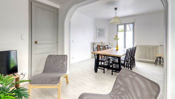 Een moderne woonkamer met een televisie, grijze fauteuils, een eettafel met stoelen en een groot raam.