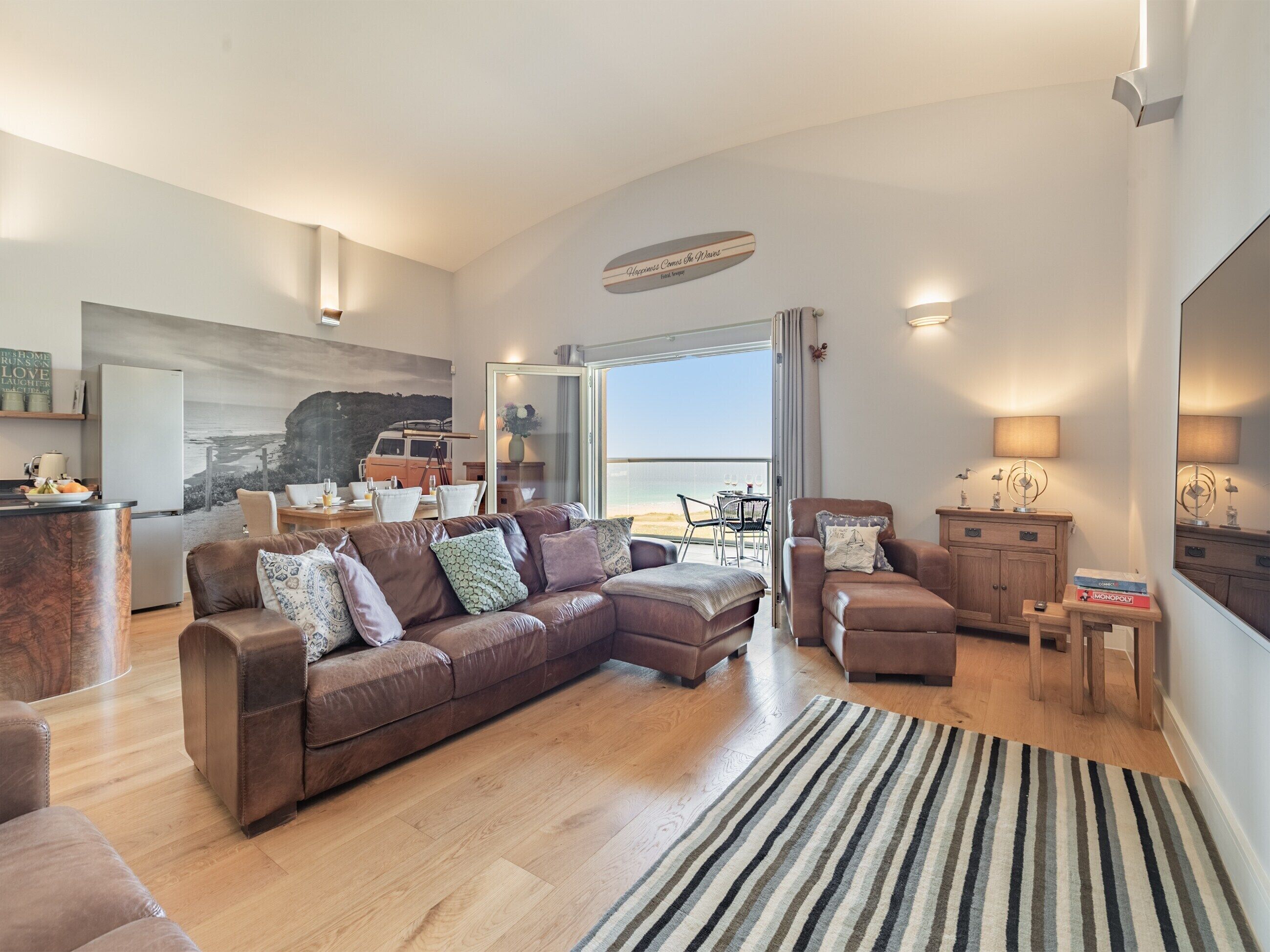 Un salon spacieux avec un grand canapé en cuir marron, un tapis à rayures et une vue sur la mer, visible à travers une porte-fenêtre coulissante.