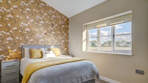 Une chambre avec un grand lit, une table de chevet, une fenêtre donnant sur un beau paysage et un papier peint à motifs floraux.