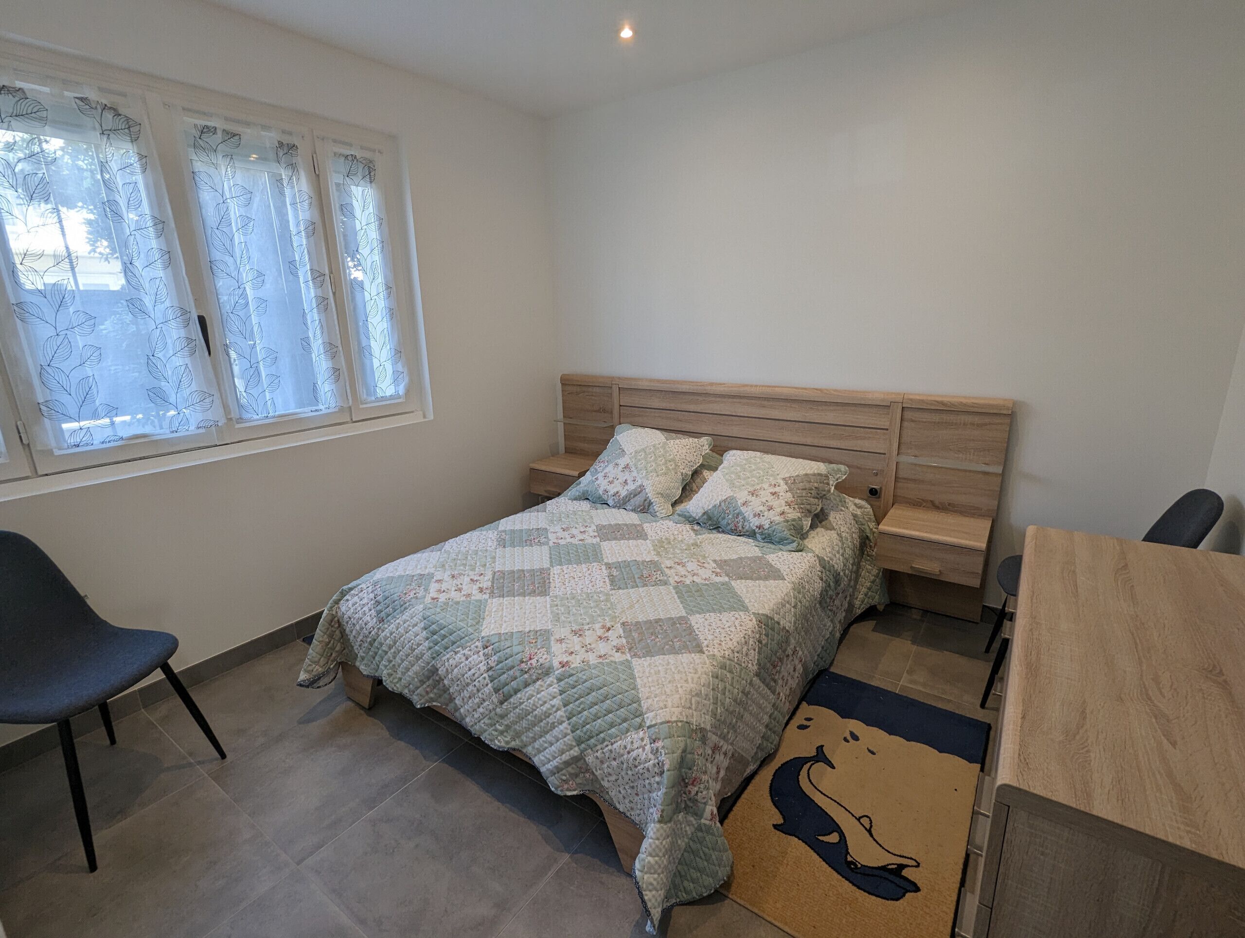 Een slaapkamer met een bed, een stoel, een nachtkastje, een houten bureau en een raam met gordijnen.