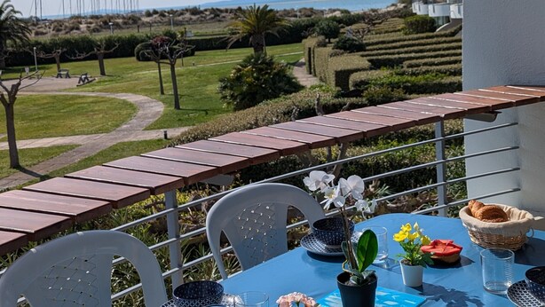 A balcony with a table set for two, overlooking a landscaped garden and a view of the sea.