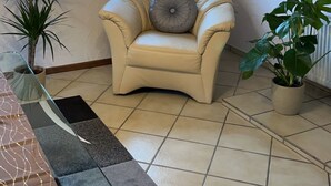 A living room with a beige leather armchair, a potted plant, a glass coffee table, a modern chandelier, and a television mounted on the wall. - Fachwerkhaus zum Hirsch in Mömbris (Mömbris)