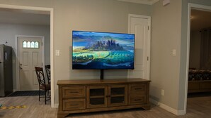 A living room with a flat-screen TV on a wooden cabinet, a door, and a chair. - Spacious Home with Luxury Finish (Brandon)