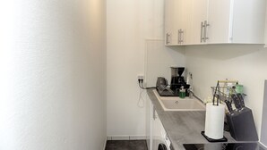 Pieni, käytännöllinen keittiö, jossa on valkoiset kaapit, kahvinkeitin, vedenkeitin ja pesuallas.