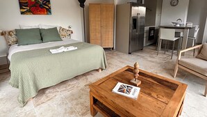 Ein Schlafzimmer mit einem Bett, einer grünen Decke, einem Holztischchen mit einer Zeitschrift, einem farbenfrohen Gemälde und einer Küchenzeile mit einem Kühlschrank und einem Esstisch.