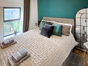 A neatly made bed with a patterned bedspread and pillows, a window with curtains, and a view of greenery outside. - Mynydd A Mor, Mountains and Sea (Trefor, near Caernarfon)