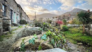 A stone building with a cobblestone pathway leading to a garden area with greenery and a small structure. - Mynydd A Mor, Mountains and Sea (Trefor, near Caernarfon)