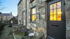 A stone building with a dark door and windows. There is a small table and chair outside. - Mynydd A Mor, Mountains and Sea (Trefor, near Caernarfon)