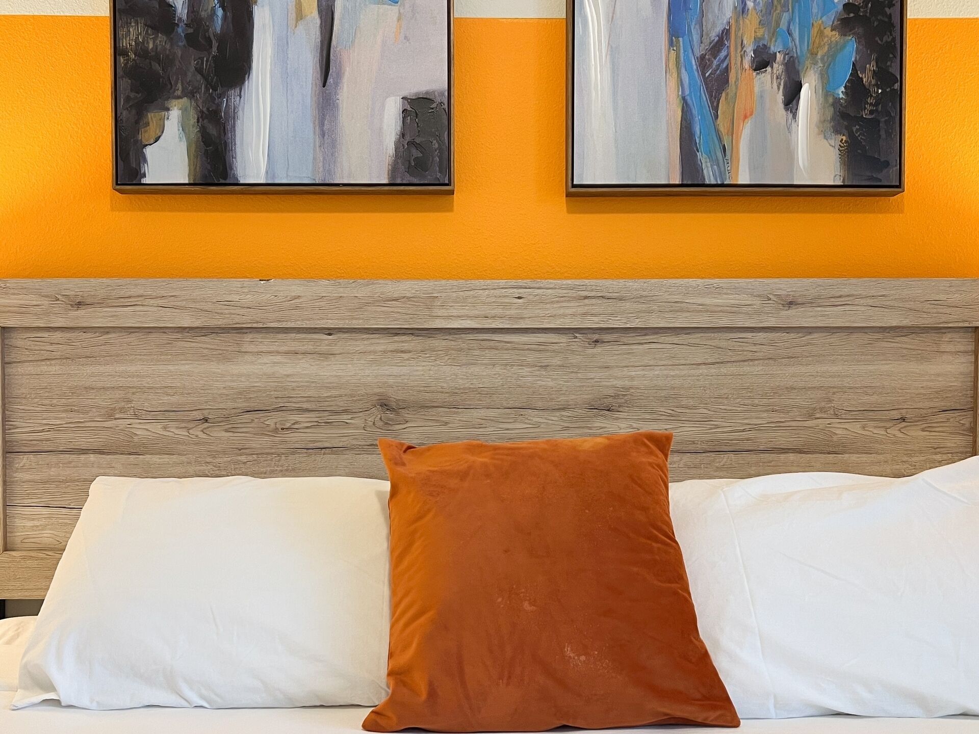 Una cama con almohadas blancas y marrones, un cabecero de madera y dos cuadros abstractos en la pared.
