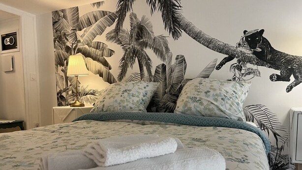 Una cama bien hecha con ropa de cama floral, un mural tropical en la pared y una mesita de noche con una lámpara.