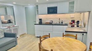 Una cocina moderna con armarios blancos, microondas y cafetera. Hay una zona de comedor con mesa y sillas de madera.