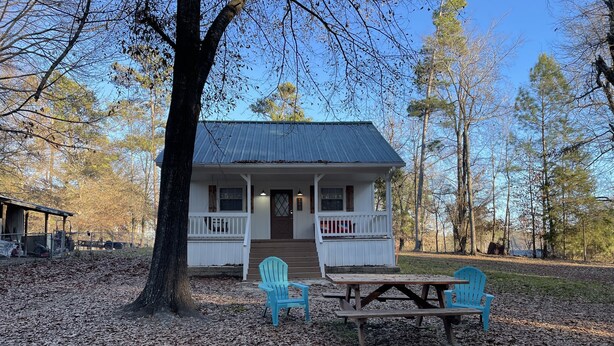Une cabane dotée d’une véranda, de deux chaises et d’une table de pique-nique, le tout entouré d’arbres et de feuilles mortes.