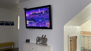 Un salon moderne avec un téléviseur à écran plat fixé au mur, un meuble blanc avec des tiroirs, un banc et un panneau indiquant « ON FACE & TIME ».