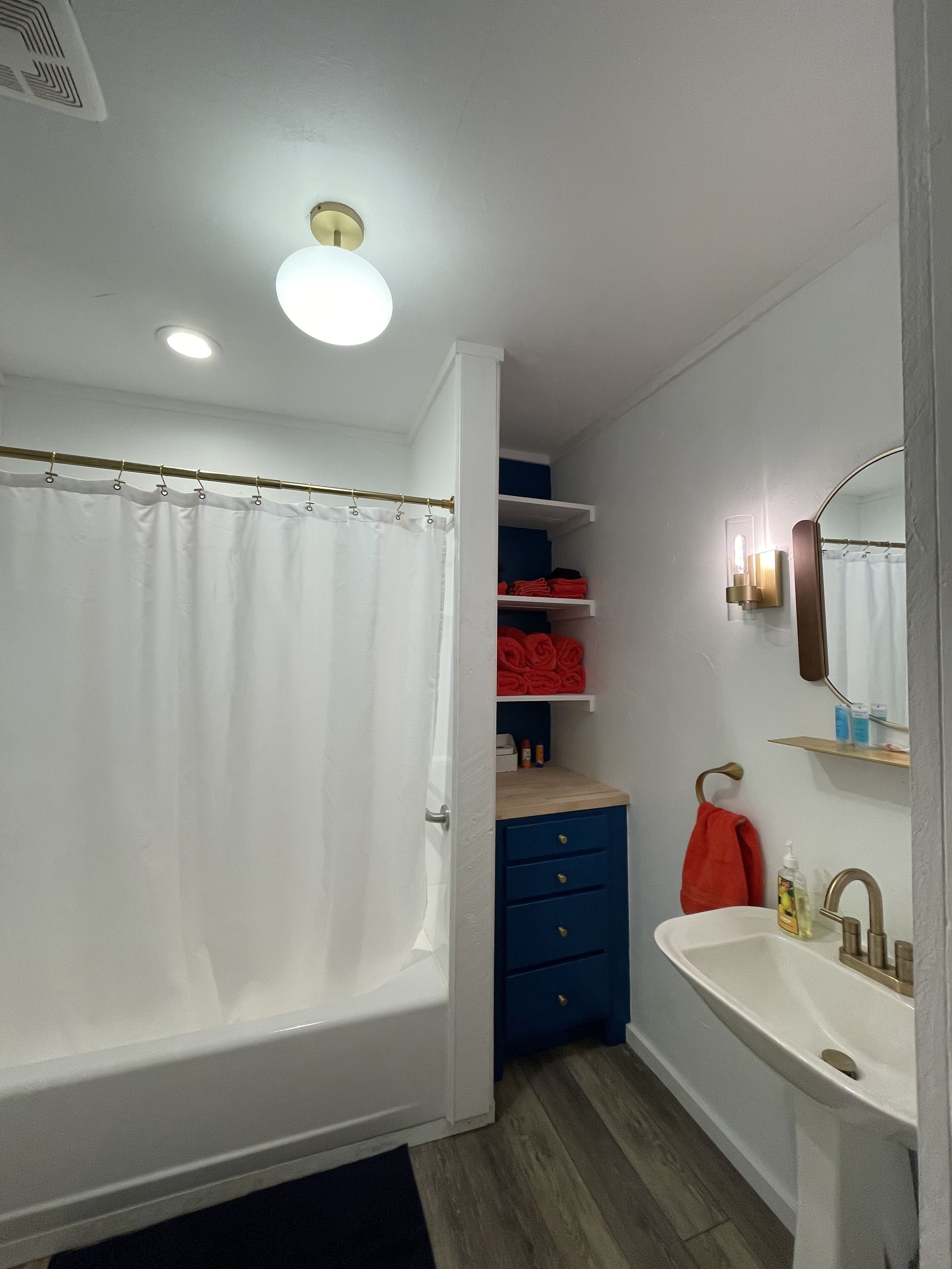 A bathroom with a bathtub, a sink, and a mirror. There are shelves with folded towels and toiletries.