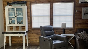 Une pièce avec un mur en bois, un fauteuil inclinable gris, un meuble blanc avec des portes en verre et une fenêtre avec des stores.