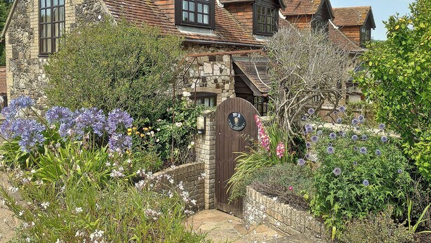 A traditional stone house with a wooden gate, surrounded by lush gardens and flowering plants. - 1 bedroom accommodation in Brighstone (Brighstone)