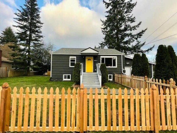 A two-story house with a yellow front door, a wooden picket fence, and a well-maintained lawn. - Spacious, stocked 1 BR Suite with Yard in Edmonds! (Edmonds)