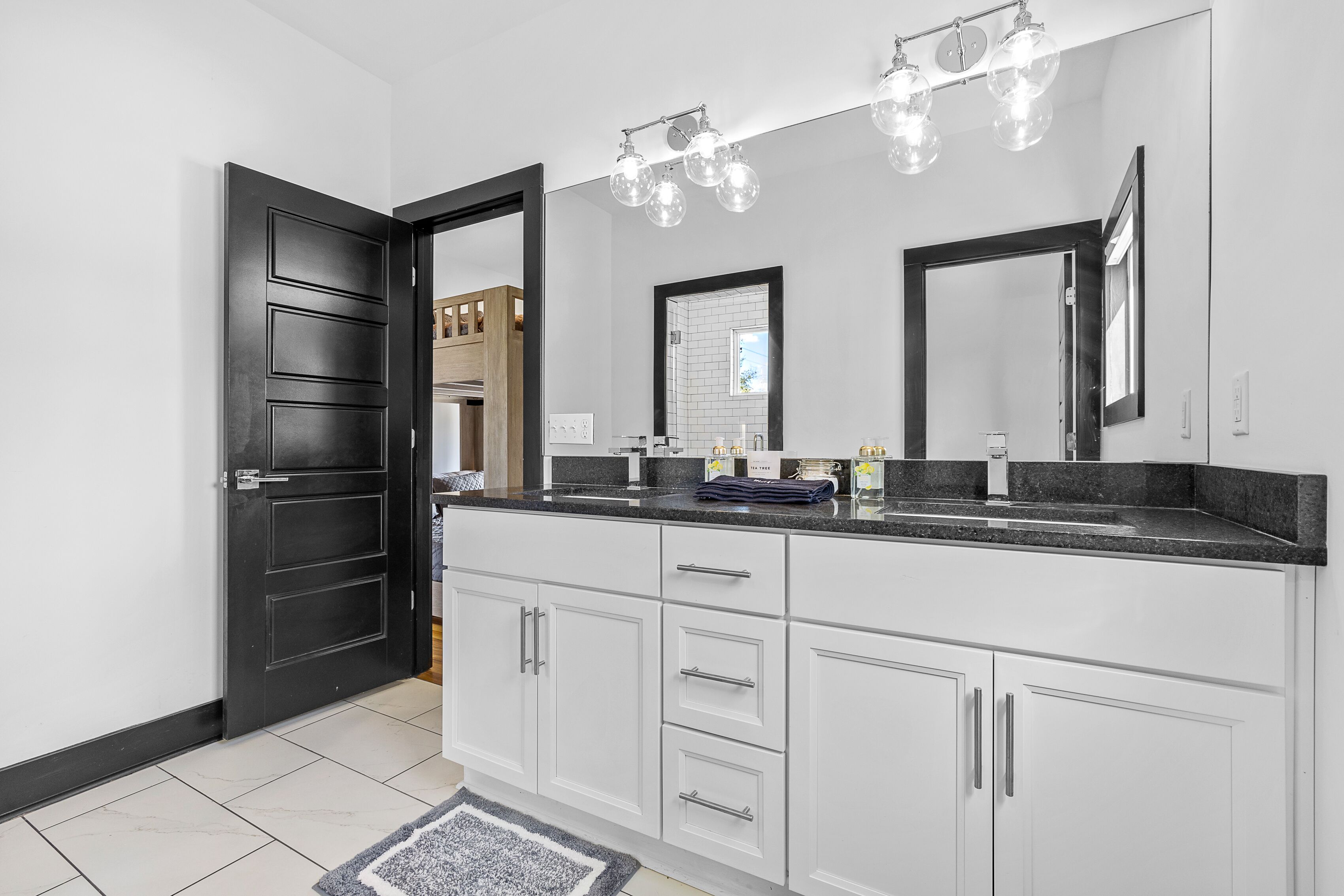 A modern bathroom with a black door, white cabinets, a granite countertop, and a large mirror with three lights.