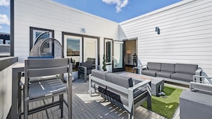 Un patio moderno al aire libre con un sofá gris, una silla de metal y una mesa pequeña.