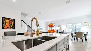 Une cuisine moderne dotée d’un grand îlot central, d’un évier en acier inoxydable et d’un robinet doré. On y trouve également un coin repas avec une table et des chaises, ainsi qu’un escalier menant à un autre niveau.