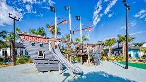 Un terrain de jeux doté d’installations ludiques sur le thème des pirates, notamment des toboggans et des structures d’escalade, le tout se dressant sur fond de palmiers et d’un ciel dégagé.