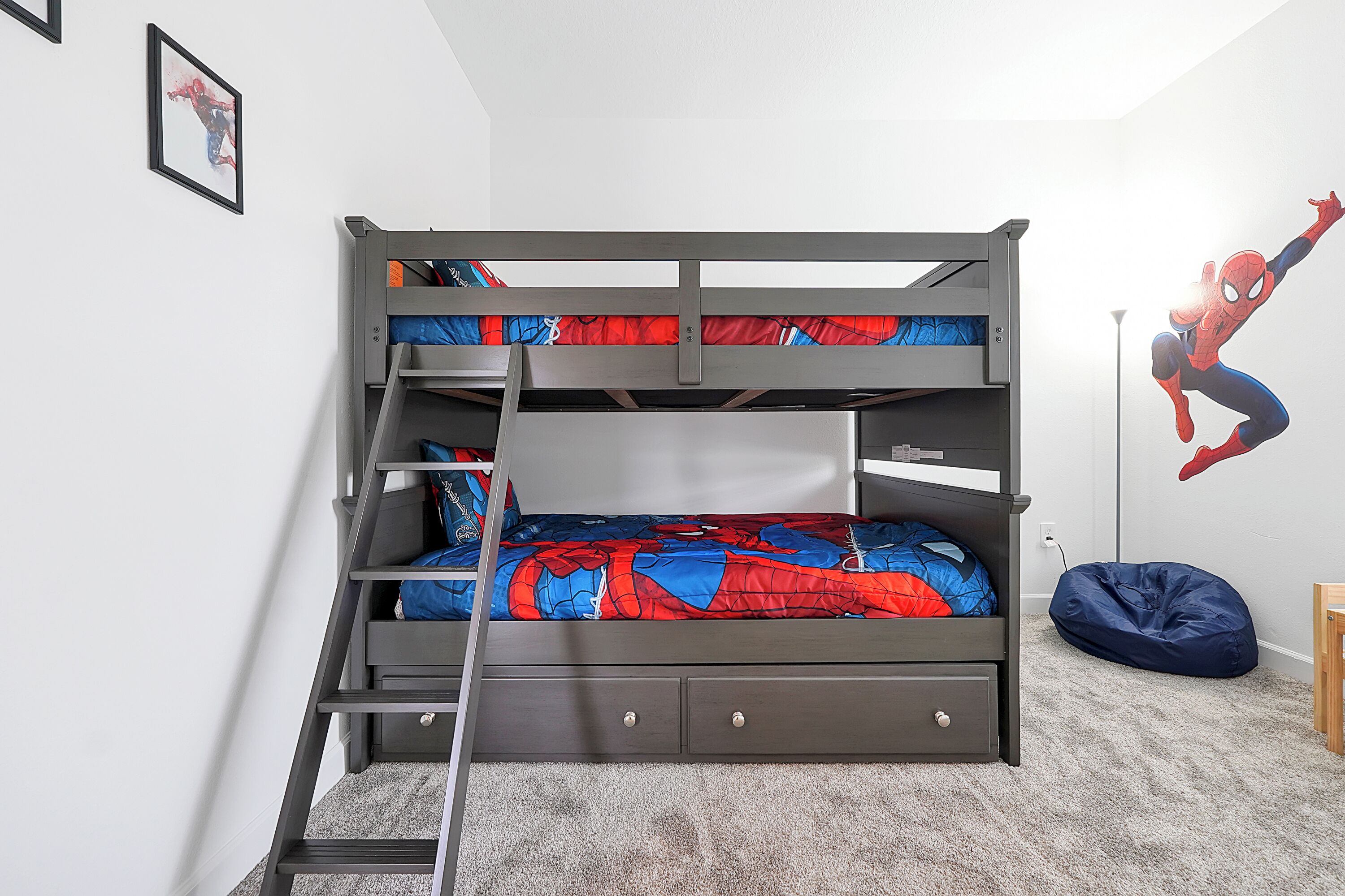 A bunk bed with a Spider-Man themed bedspread, a ladder, and a bean bag chair in a room with a carpeted floor and a wall-mounted lamp.