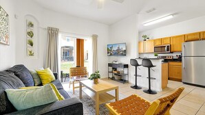 Un appartement moderne comprenant un salon, un coin repas et une cuisine. Il est équipé d'un canapé, d'une table, de deux tabourets de bar, d'un téléviseur, de meubles de rangement en bois, d'un four à micro-ondes et d'un réfrigérateur.