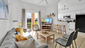 En moderne stue med sofa, sofabord og fjernsyn, et køkken med hvide skabe og hårde hvidevarer, samt en balkon med gardiner.