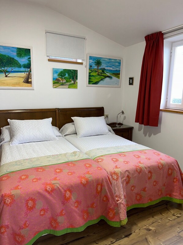 A double bed with white bedding and a pink floral bedspread, two white pillows, a wooden headboard, and a nightstand with a lamp.