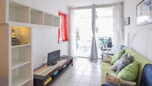 A living room with a TV on a shelf, a green sofa with cushions, a red curtain, and a view through a glass door to an outdoor area. - Cozy apartment in Isca Marina with WiFi (Isca Marina)