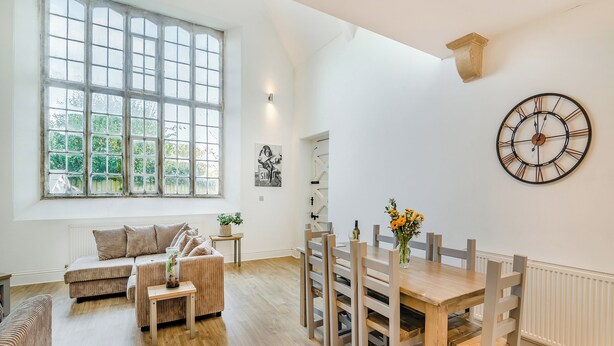 Ein geräumiger Raum mit großem Fenster, einem Esstisch mit Stühlen, einer Couch und einer Wanduhr.