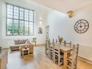 A spacious room with a large window, a dining table with chairs, a sofa, and a wall-mounted clock.