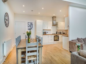 A modern kitchen with white cabinets, a dining area with a wooden table and chairs, and a living area with striped sofas.
