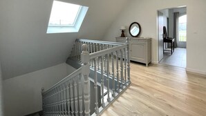 Una habitación amplia con techo alto, un tragaluz, una escalera con balaustres blancos, suelo de madera y vistas al exterior a través de una puerta.
