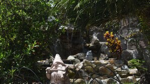Un jardín con una estatua de una figura sosteniendo un báculo, rodeado de exuberante vegetación y una pequeña cascada.