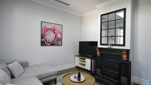 A living room with a grey sofa, a flat-screen TV, a fireplace, and a large floral painting on the wall. - Beautifully renovated country cottage located near Mclaren Vale's best wineries (Clarendon)
