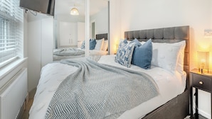 Un dormitorio moderno con una cama grande, un televisor montado en la pared, un espejo y una mesita de noche con una lámpara.