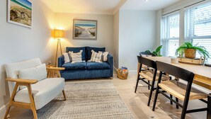 Una sala de estar acogedora con un sofá azul, un sillón blanco, una mesa de centro de madera y una mesa de comedor con sillas. Hay una lámpara, un cuadro enmarcado en la pared y una ventana con persianas.