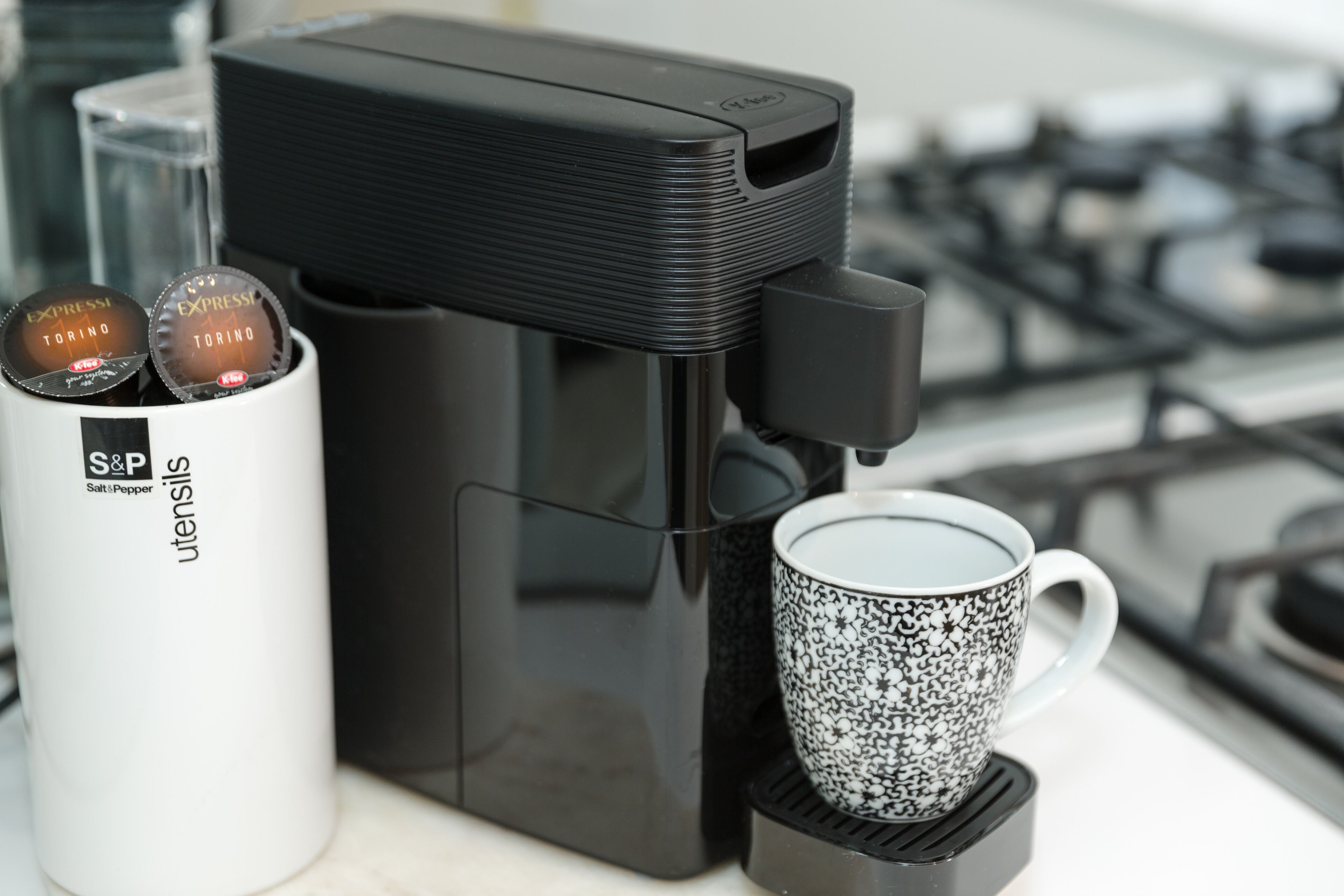 Une machine à café avec une tasse blanche posée sur un sous-verre noir, un récipient blanc portant les inscriptions « S&P » et « ustensiles », et un arrière-plan flou avec une cuisinière.