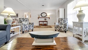 Ein Wohnzimmer mit einem großen, blauen Mittelstück in Form einer Schale, zwei gemusterten Sesseln, einem Sofa und einem weißen Beistelltisch mit Lampe.