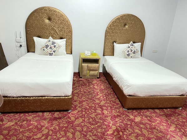 A hotel room with two beds, each with a headboard, white bedding, and patterned pillows. There is a small bedside table with a phone and a lamp. The room has a red patterned carpet. - Royal Beach Hotel (Shinas)