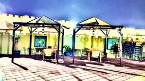 Two outdoor seating areas with tables and chairs under pergolas, set against a wall with a mural. - Royal Beach Hotel (Shinas)