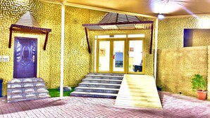 A building entrance with a stone wall, a wooden door, and a set of stairs leading to a covered porch area. - Royal Beach Hotel (Shinas)