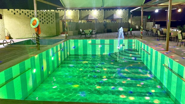 An outdoor swimming pool with a green-tiled edge, a water fountain, and a covered seating area with chairs and tables. - Royal Beach Hotel (Shinas)