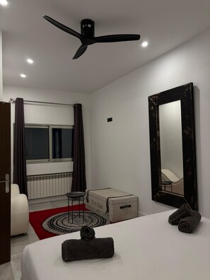 Une chambre moderne comprenant un lit, un grand miroir, un ventilateur de plafond, une fenêtre avec des rideaux, un radiateur et une petite table avec une chaise.