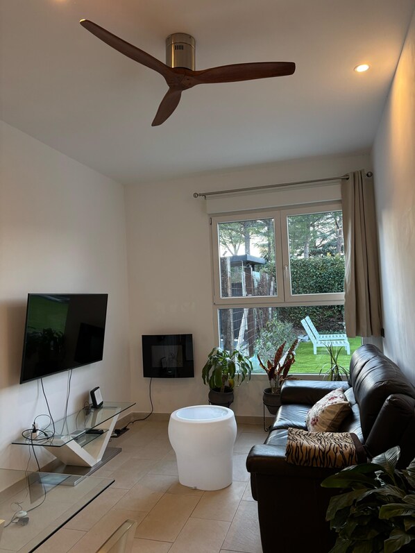 Un salon moderne doté d’un ventilateur de plafond, d’un téléviseur à écran plat, d’une table basse en verre, d’un canapé en cuir et offrant une vue sur un jardin grâce à de grandes fenêtres.