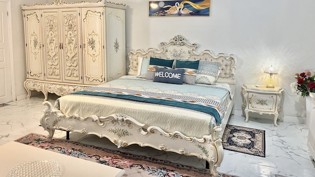 Ein Schlafzimmer mit einem großen, verzierten Bett, einem passenden Kleiderschrank, einem kleinen Nachttisch mit Lampe und einem floralen Teppich.